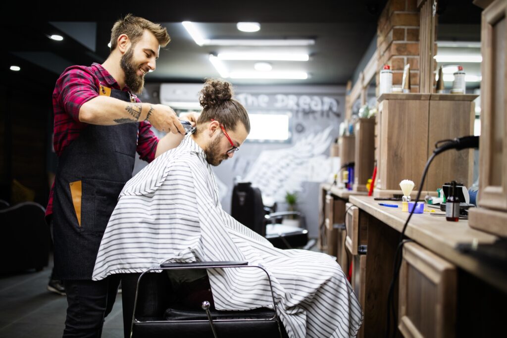 Young bearded man getting haircut by hairdresser while sitting in chair at barbershop salon