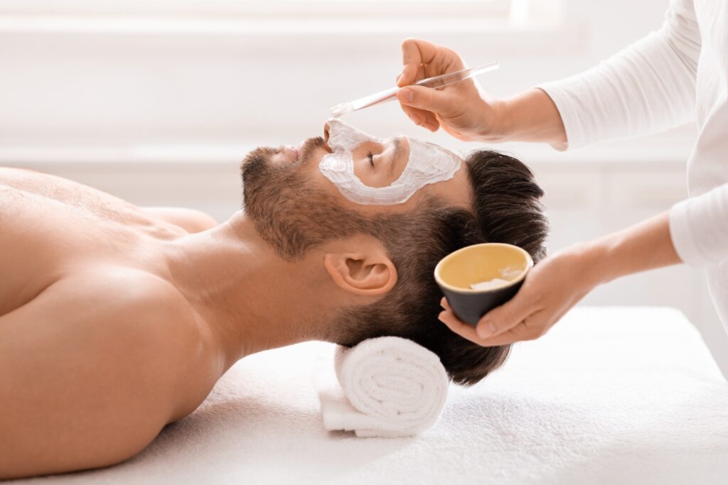 Side view of bearded man getting face treatment at modern spa salon. Female attendant applying nourishing face mask on sleeping middle aged man face. Male cosmetology and face care concept